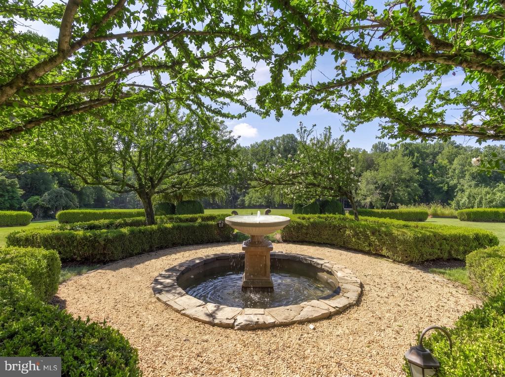 Landscaped garden with a central stone fountain, gravel, clipped hedges, and mature trees.