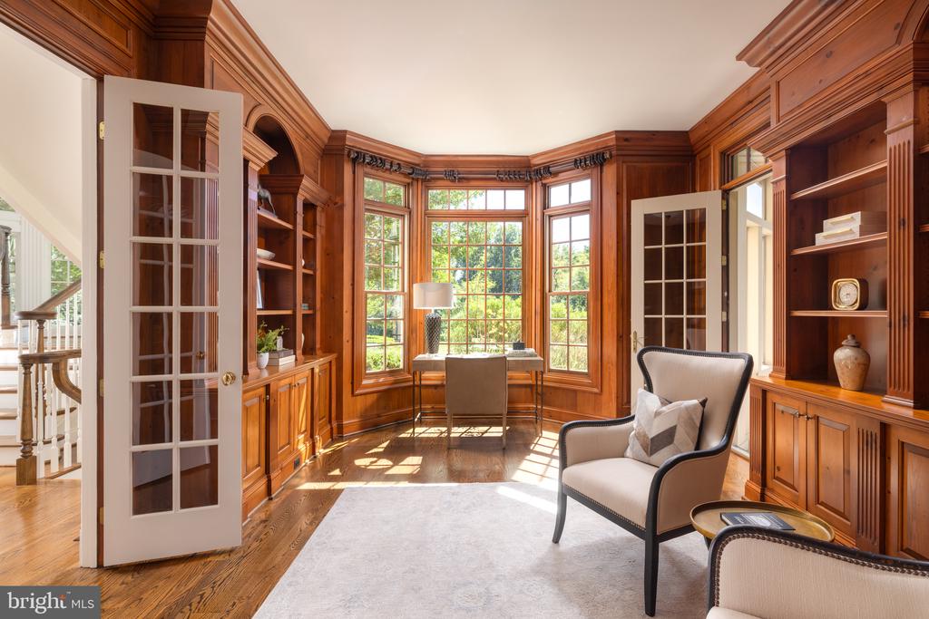 Wood-paneled study with built-in shelves, a sunlit bay window, desk, and upholstered chairs.