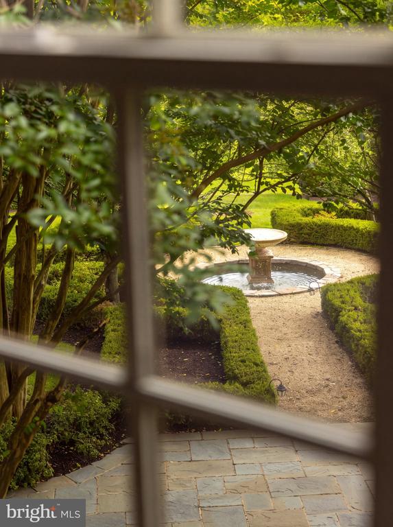 Landscaped garden with a stone fountain, clipped hedges, and gravel path, viewed through a window.