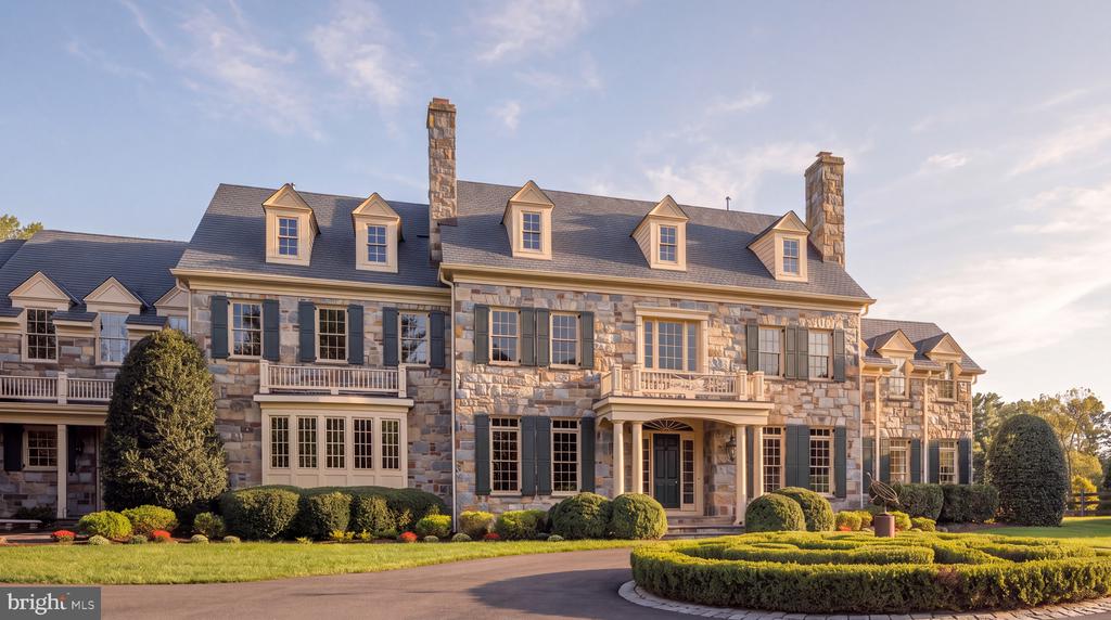 Large stone home with dormer windows, two chimneys, shaped hedges, and a curved driveway.