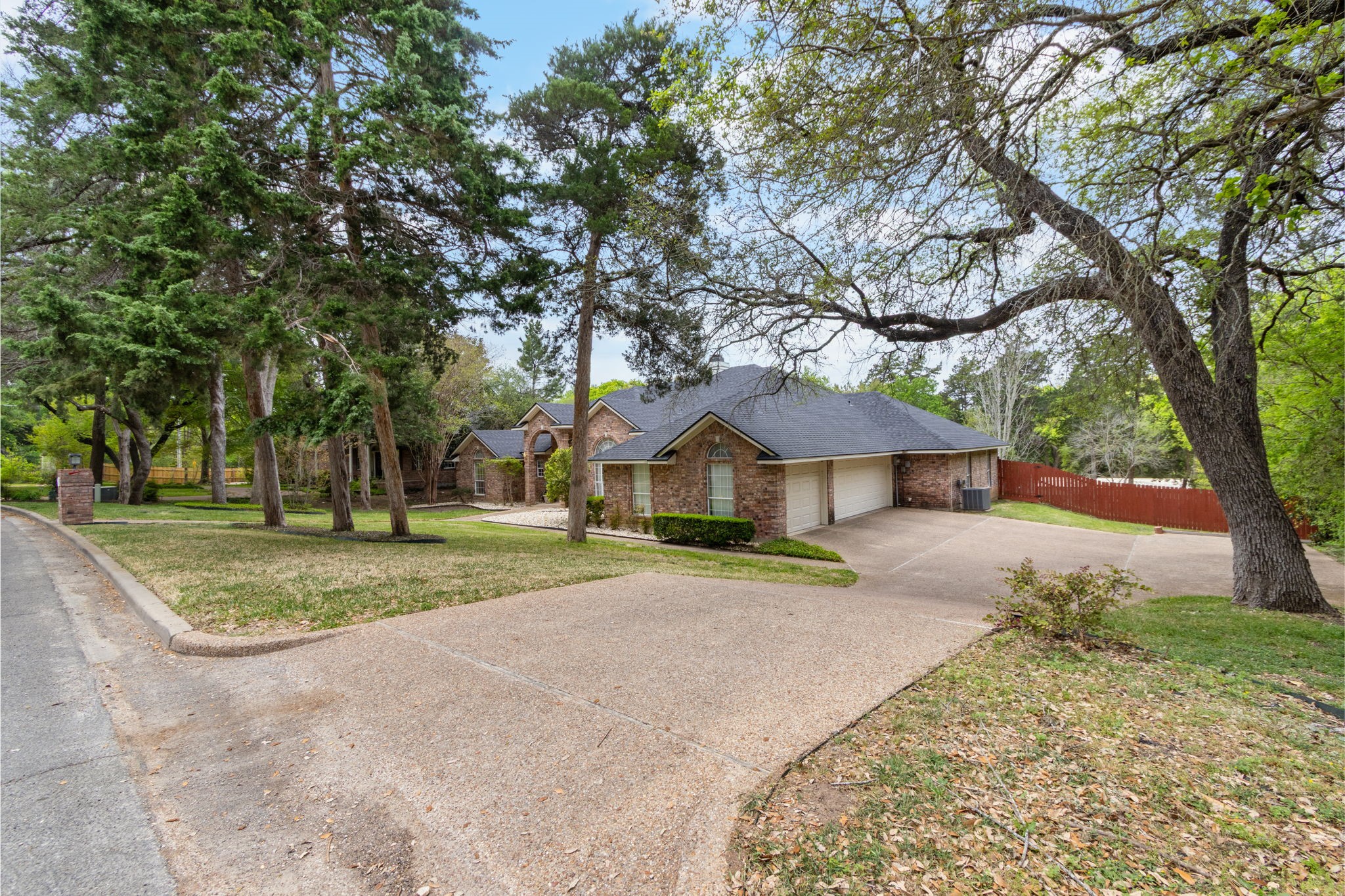 Brick home with a two-car garage, broad driveway, mature trees, and a red side-yard fence.