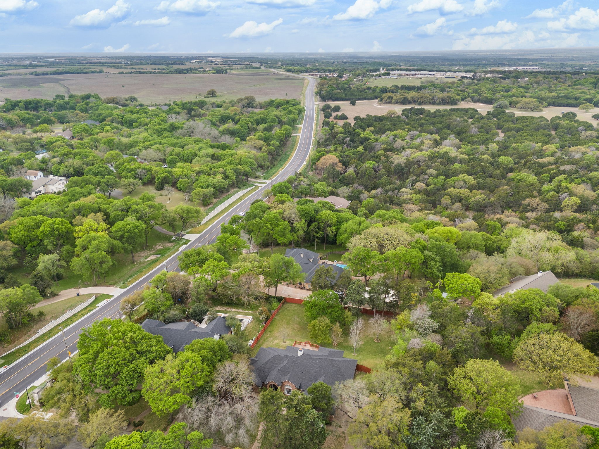 Aerial view of a tree-surrounded home beside a curving road and neighboring properties.