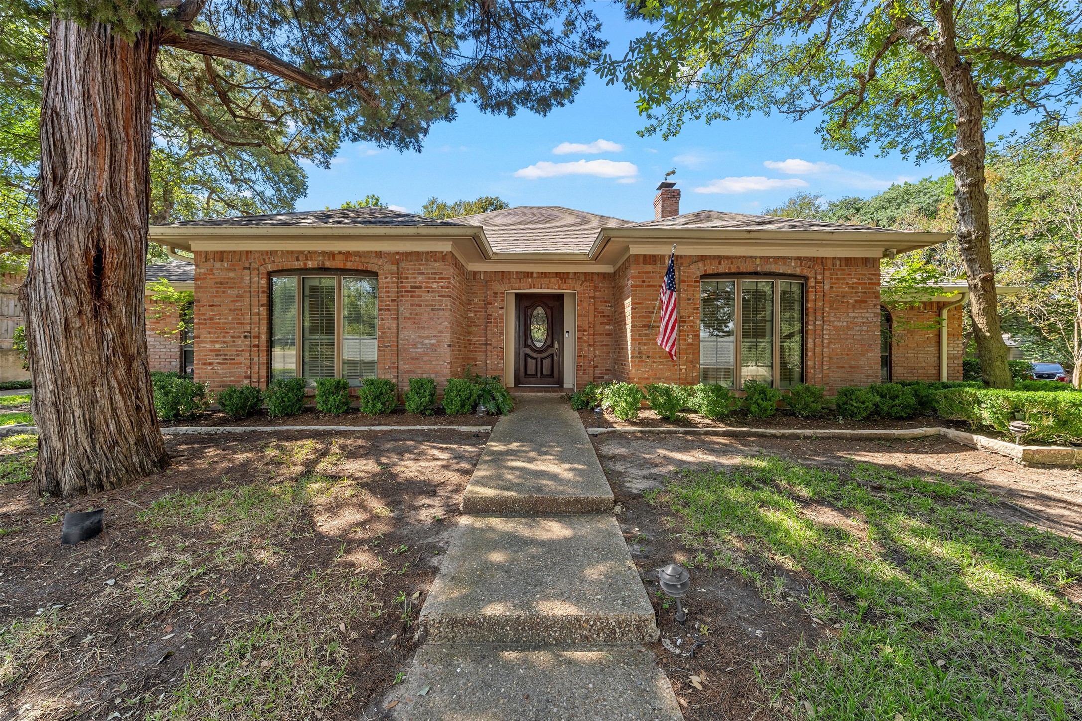 Red brick house with an American flag, concrete walkway, and mature shade trees.
