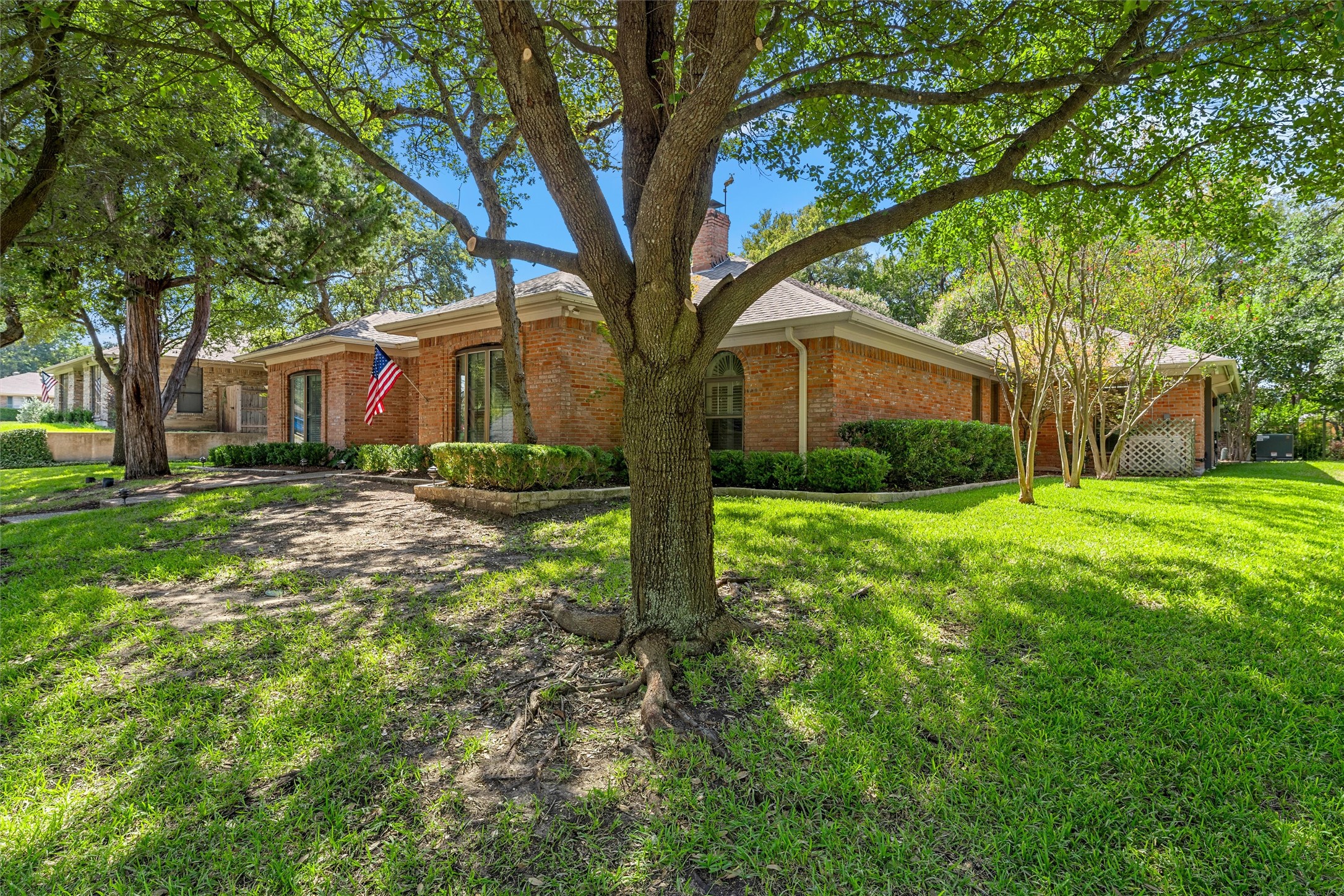 Red brick house partly shaded by a large mature tree, with a green lawn and American flag.
