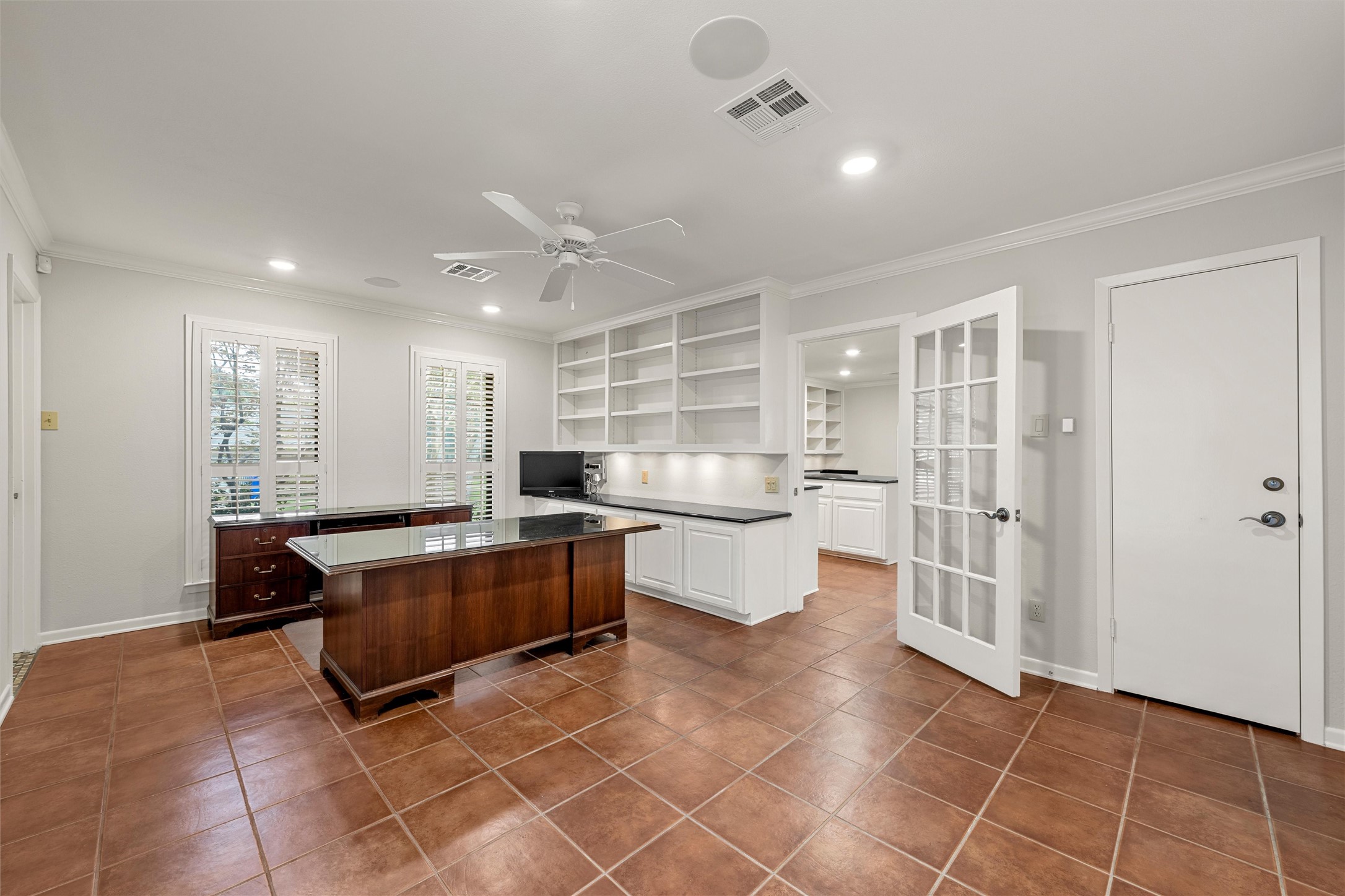 Home office with built-in shelving, a dark wood desk, shuttered windows, and terracotta tile flooring.