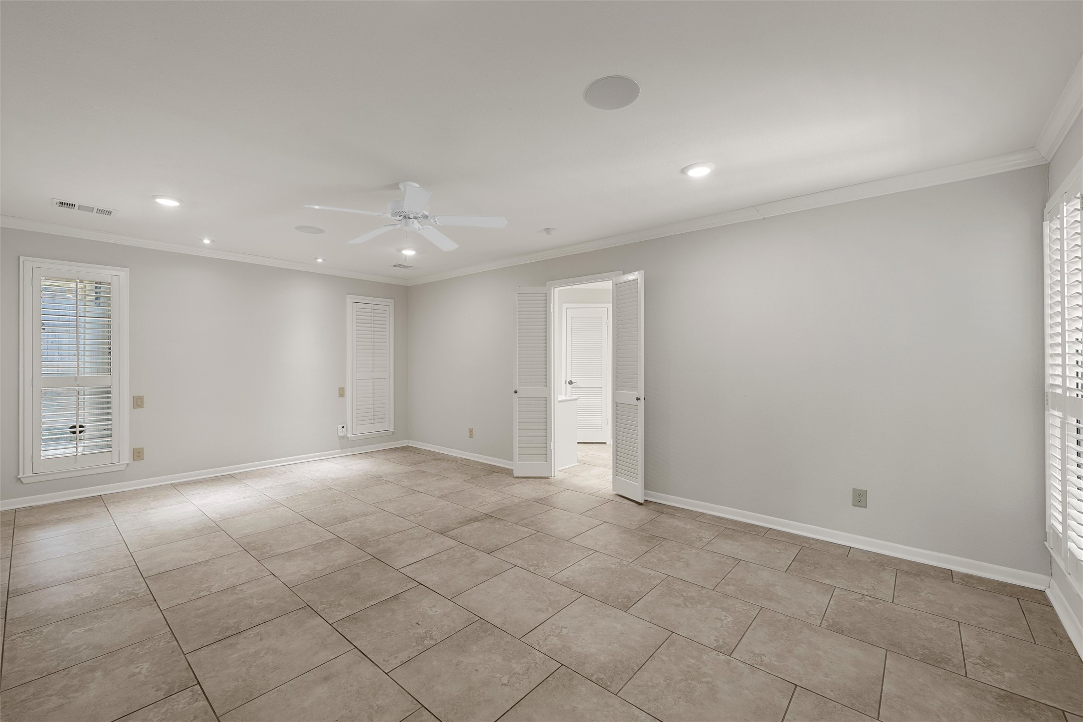 Empty room with large beige tile floors, plantation-shuttered windows, and an open louvered doorway.