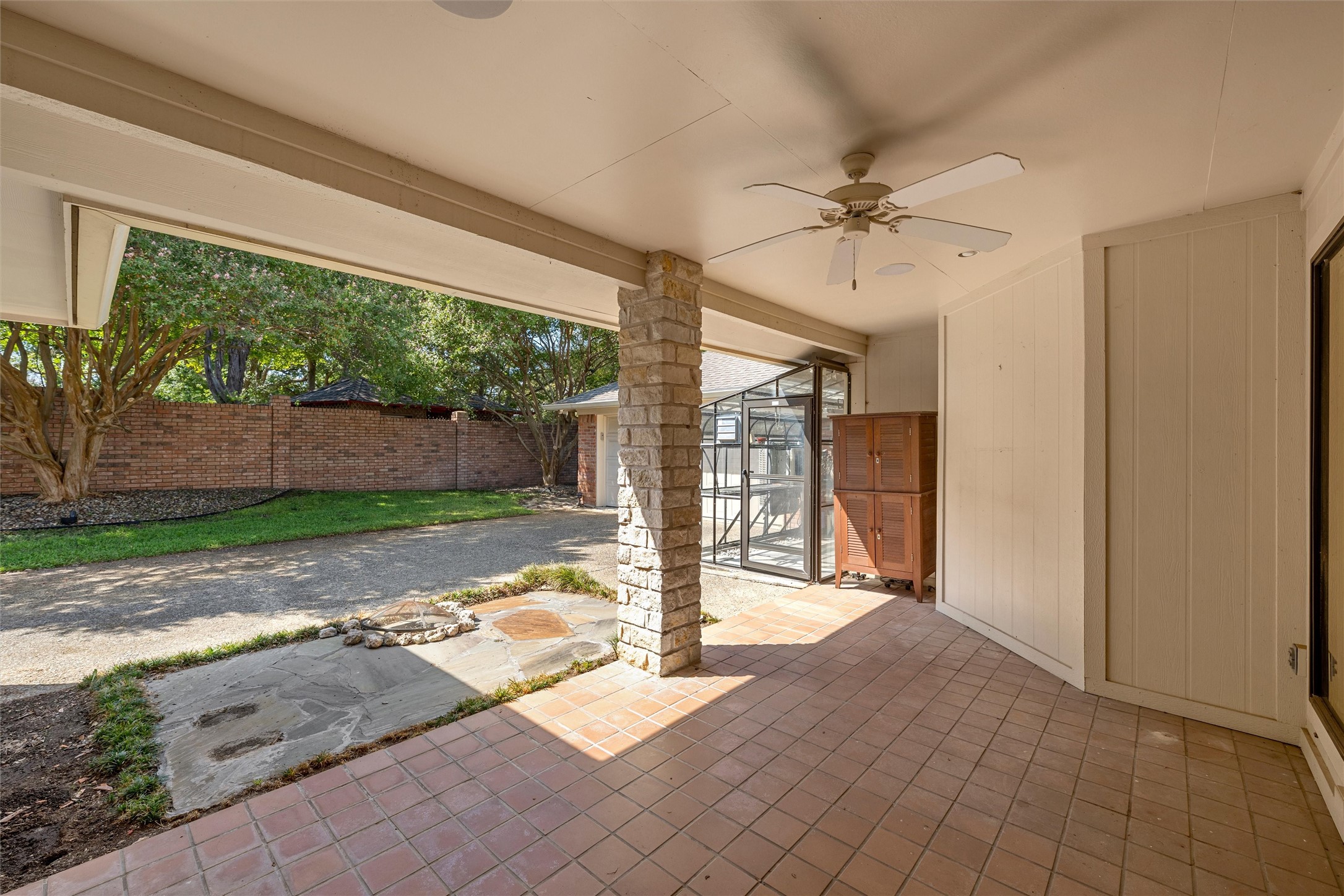 Covered patio with terracotta tile, ceiling fan, and stone column overlooking a fenced, tree-lined yard.