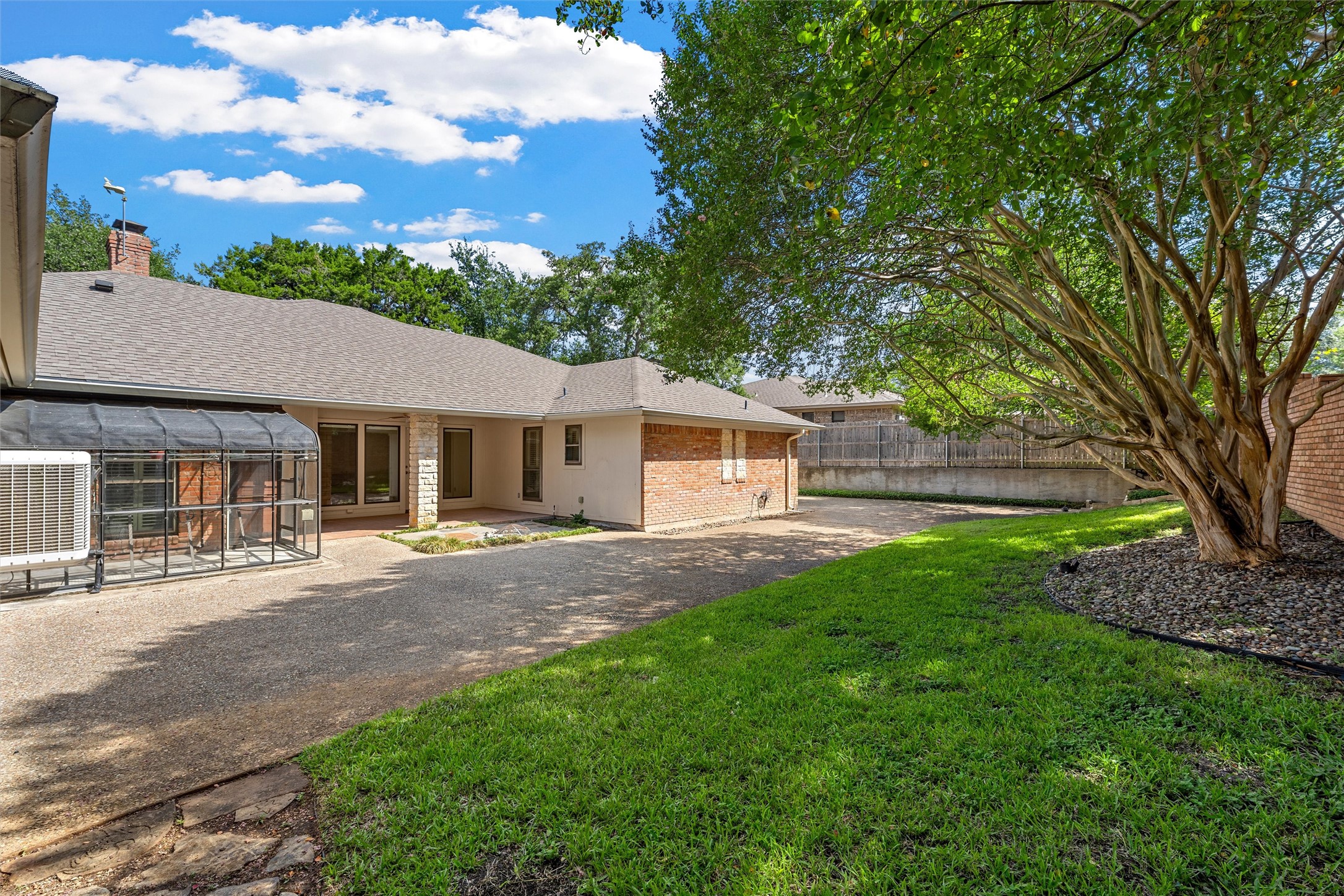 Home’s rear exterior with a covered patio, screened enclosure, green lawn, and sprawling shade tree.