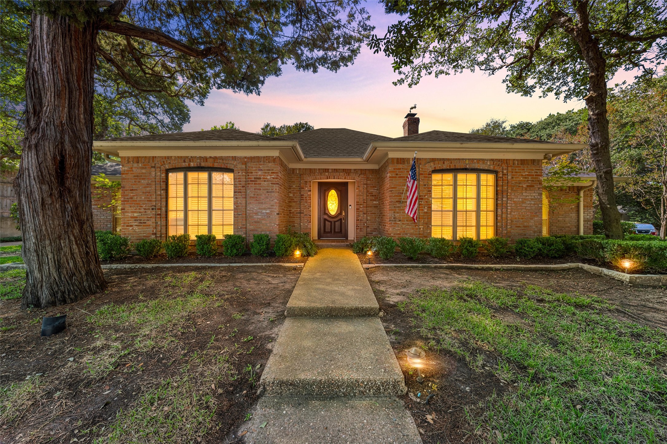 Red-brick home framed by mature trees, with a front walkway and warmly lit windows at dusk.