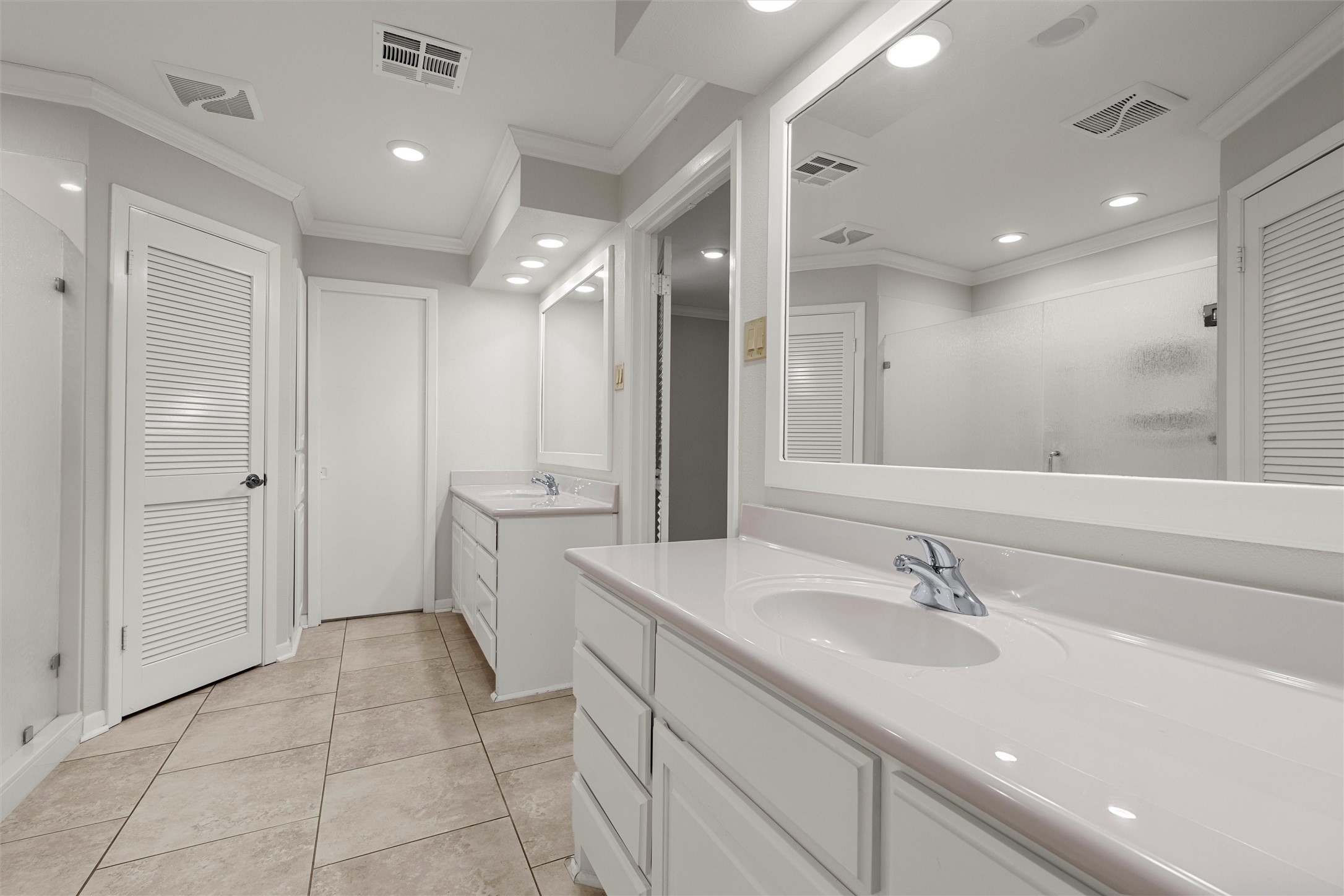 Bathroom with two white vanities, large mirrors, and beige tiled flooring.