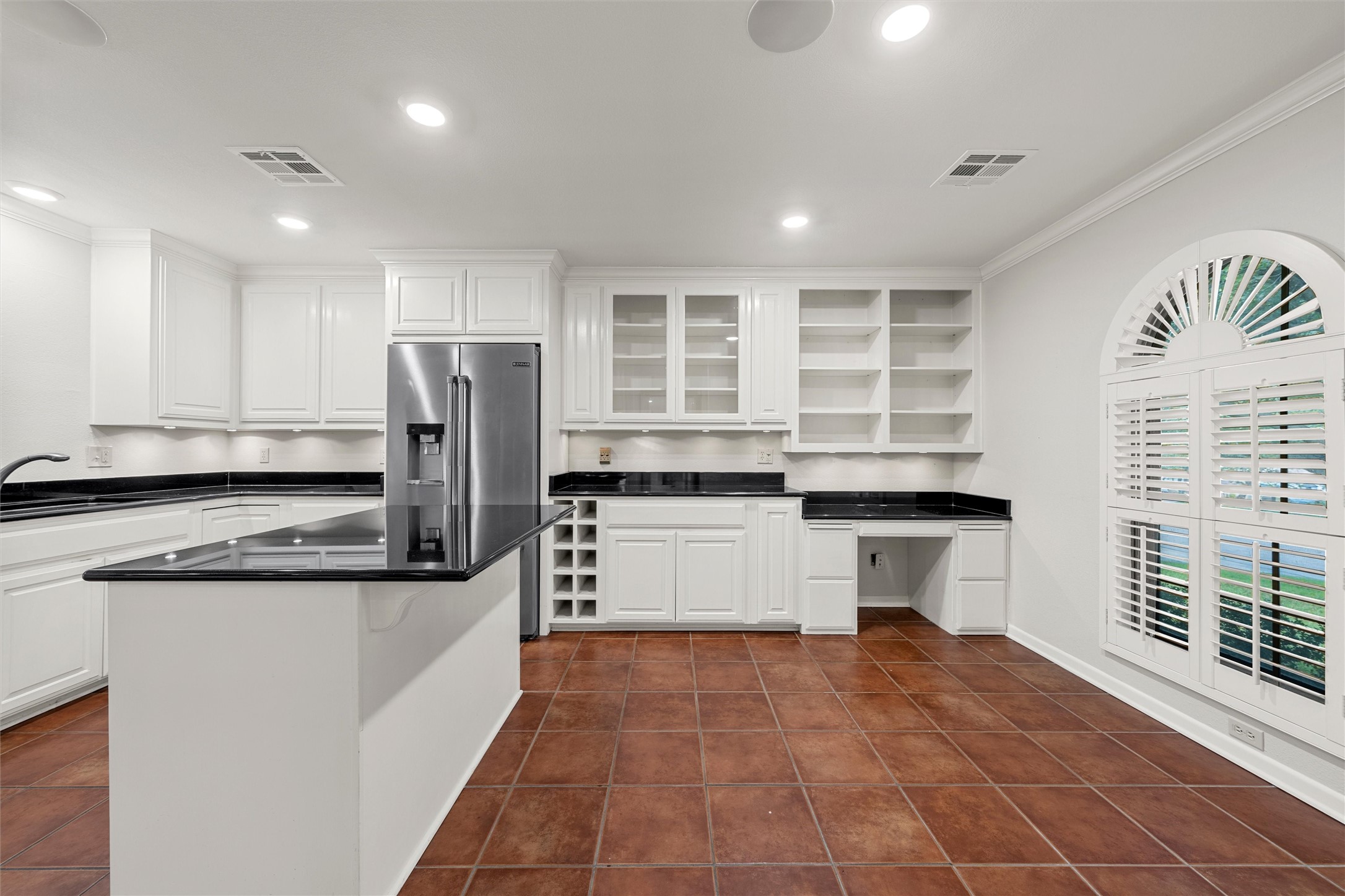 Kitchen with white cabinetry, black counters, an island, and arched plantation shutters.