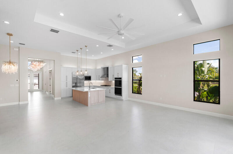 Vacant open-plan kitchen with a wood-paneled island, white cabinets, and windows overlooking tropical greenery.