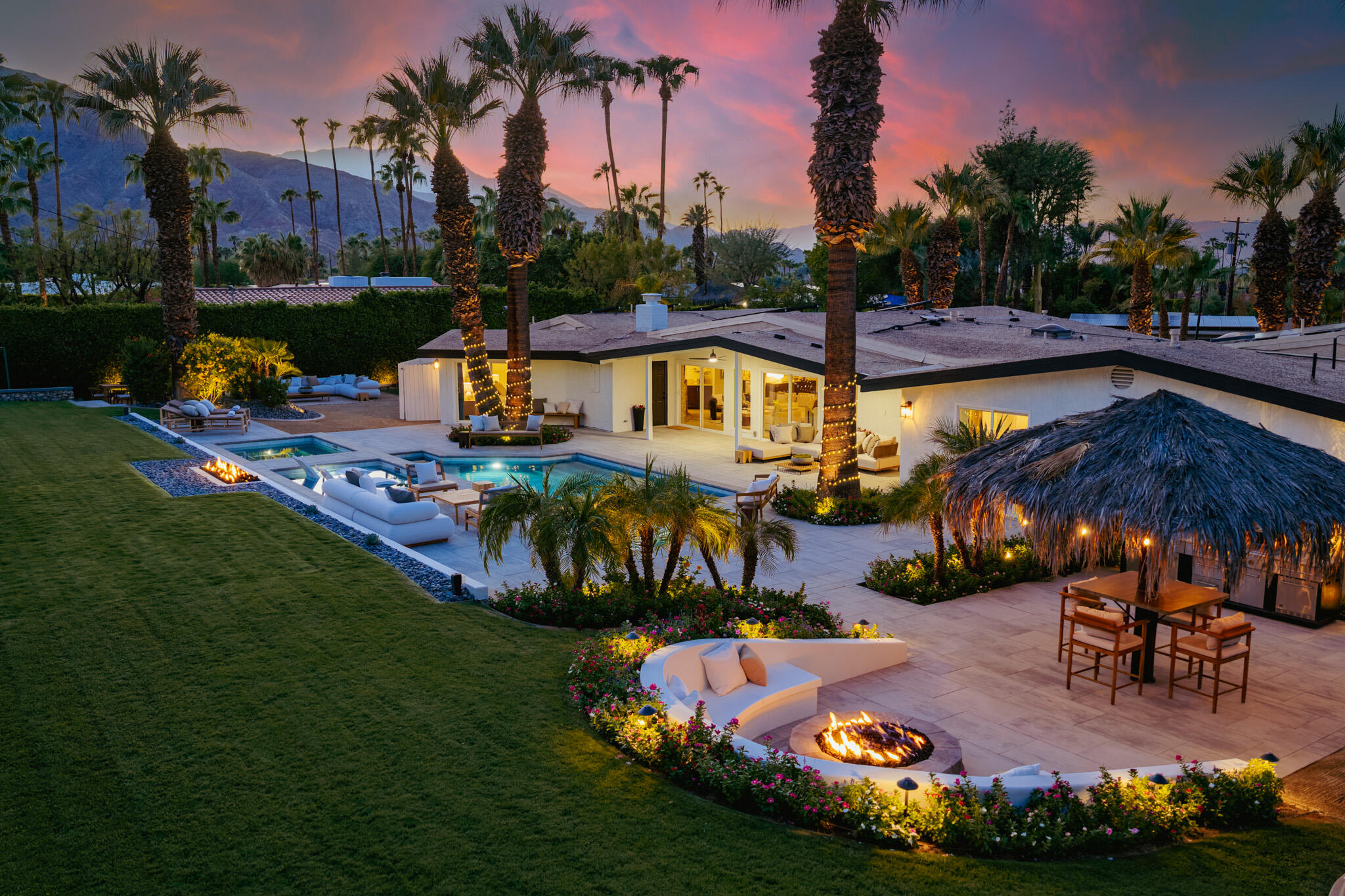 Modern home with a pool, palm-lined patio, fire pits, and mountain views at dusk.