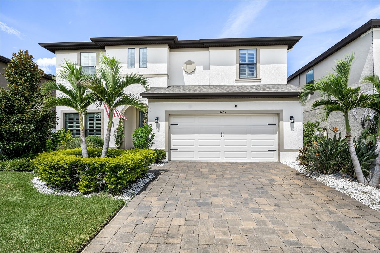 Two-story white stucco home with palm trees, a double garage, and a wide paver driveway.