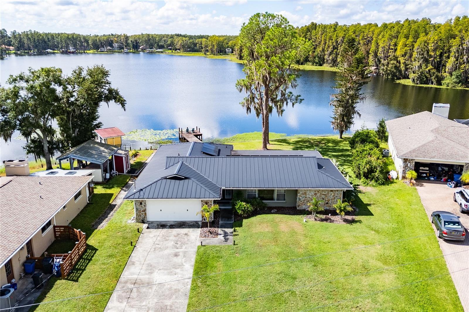 Aerial view of a lakefront home with a dark metal roof, grassy yard, and small dock.