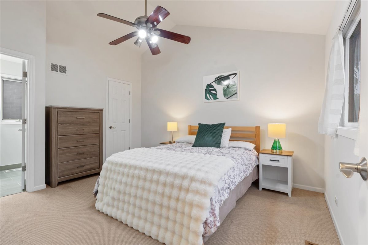 Furnished bedroom with a large white bed, wooden dresser, ceiling fan, and doorway to a tiled bathroom.