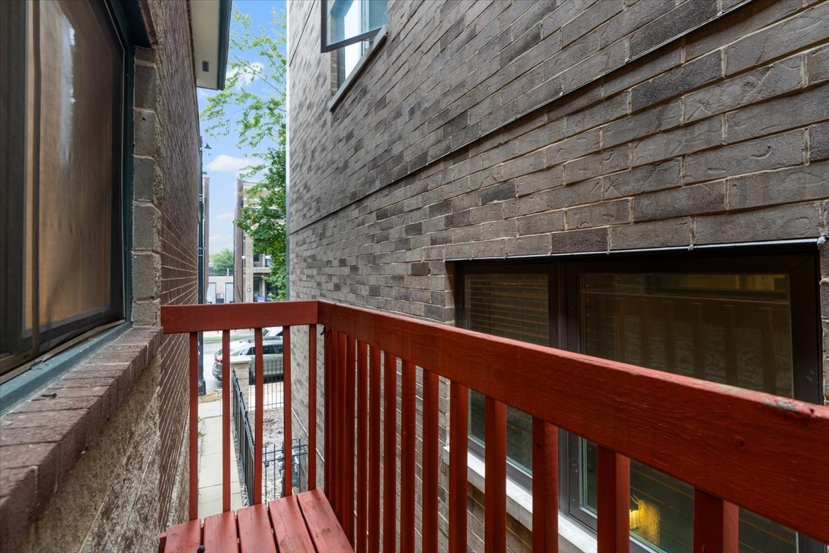 Narrow brick-walled balcony with red wooden railing overlooking a leafy street.