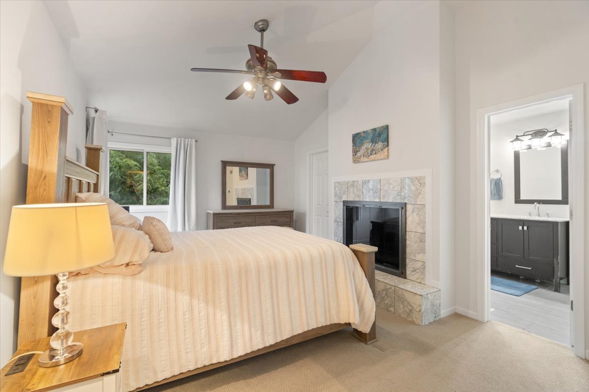 Furnished bedroom with a tiled fireplace, ceiling fan, and adjoining bathroom.