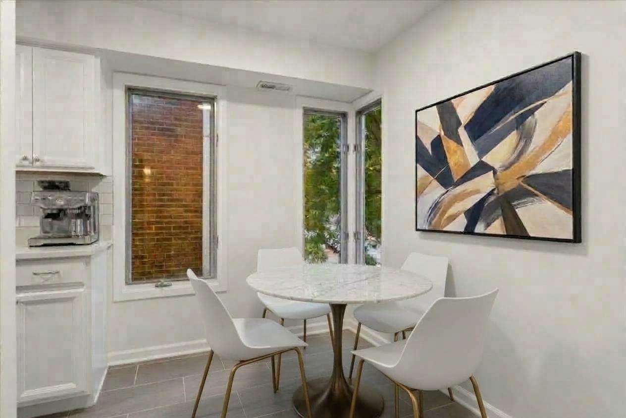 Dining nook with a round table and white chairs beside corner windows and kitchen cabinetry.