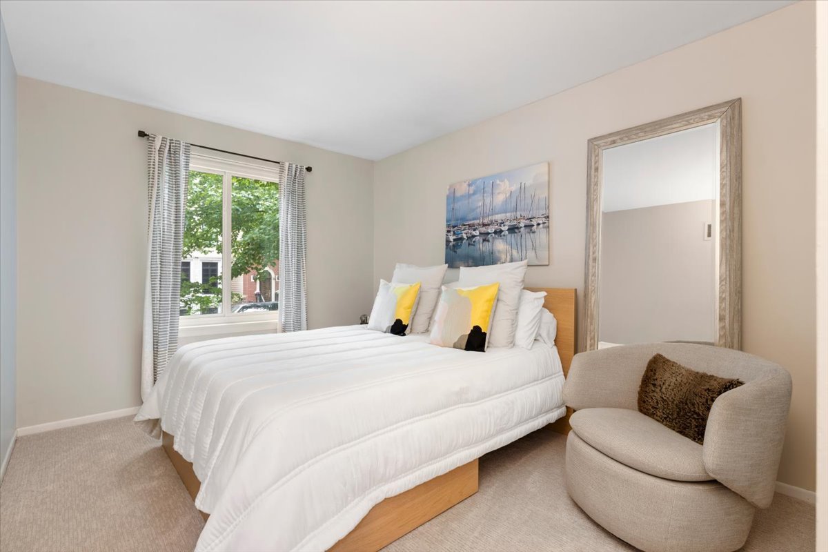 Furnished bedroom with a white bed, yellow accent pillows, and a rounded chair beside a full-length mirror.