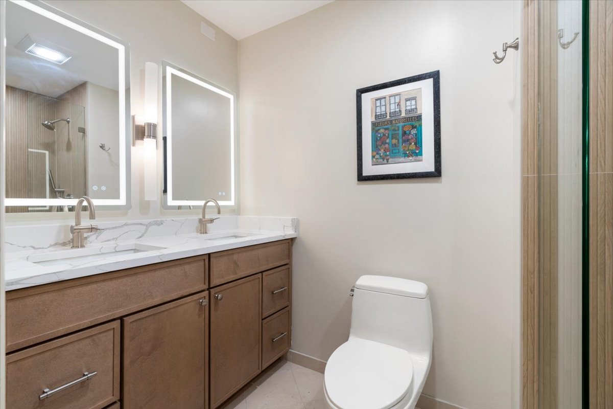 Bathroom with a double-sink wood vanity, illuminated mirrors, and a glass-enclosed shower.