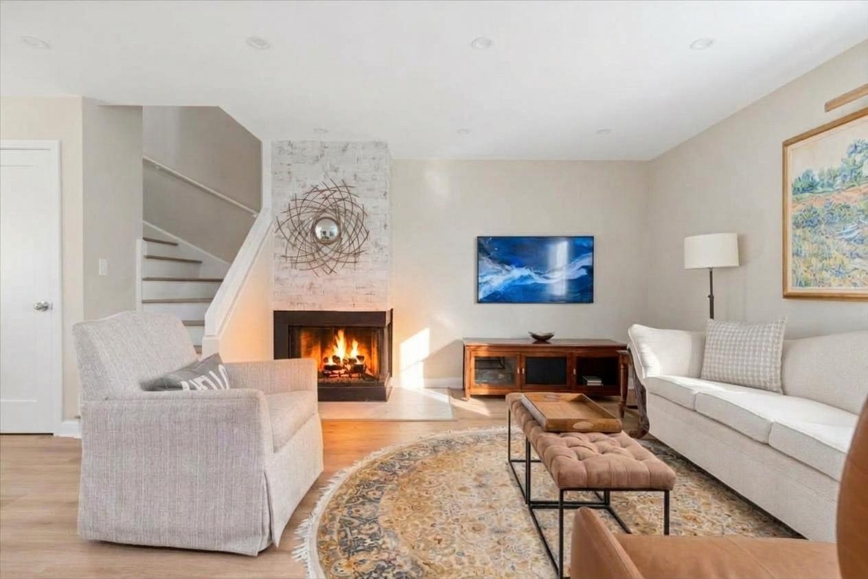 Living room with a lit fireplace, staircase, pale sofas, and patterned rug.