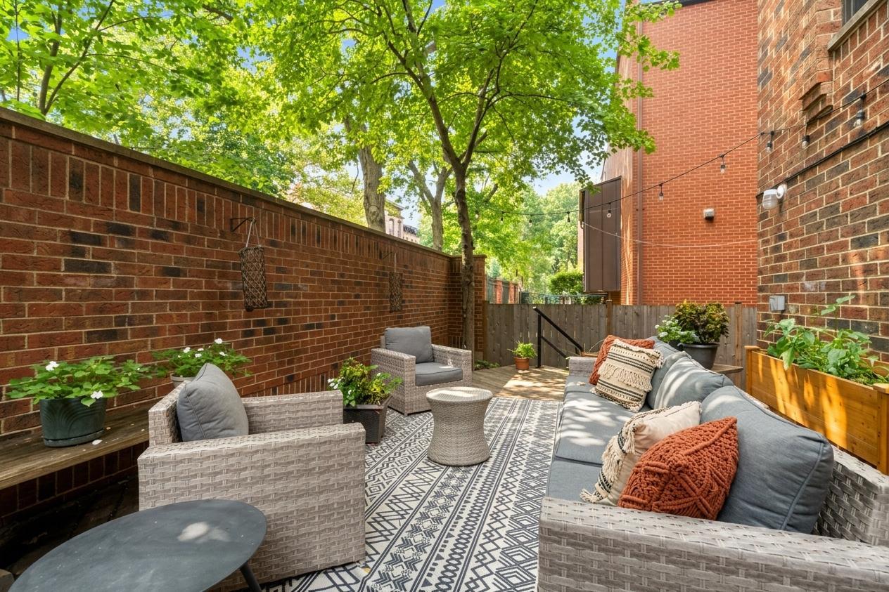 Furnished brick-walled patio with wicker seating, patterned rugs, and leafy trees overhead.