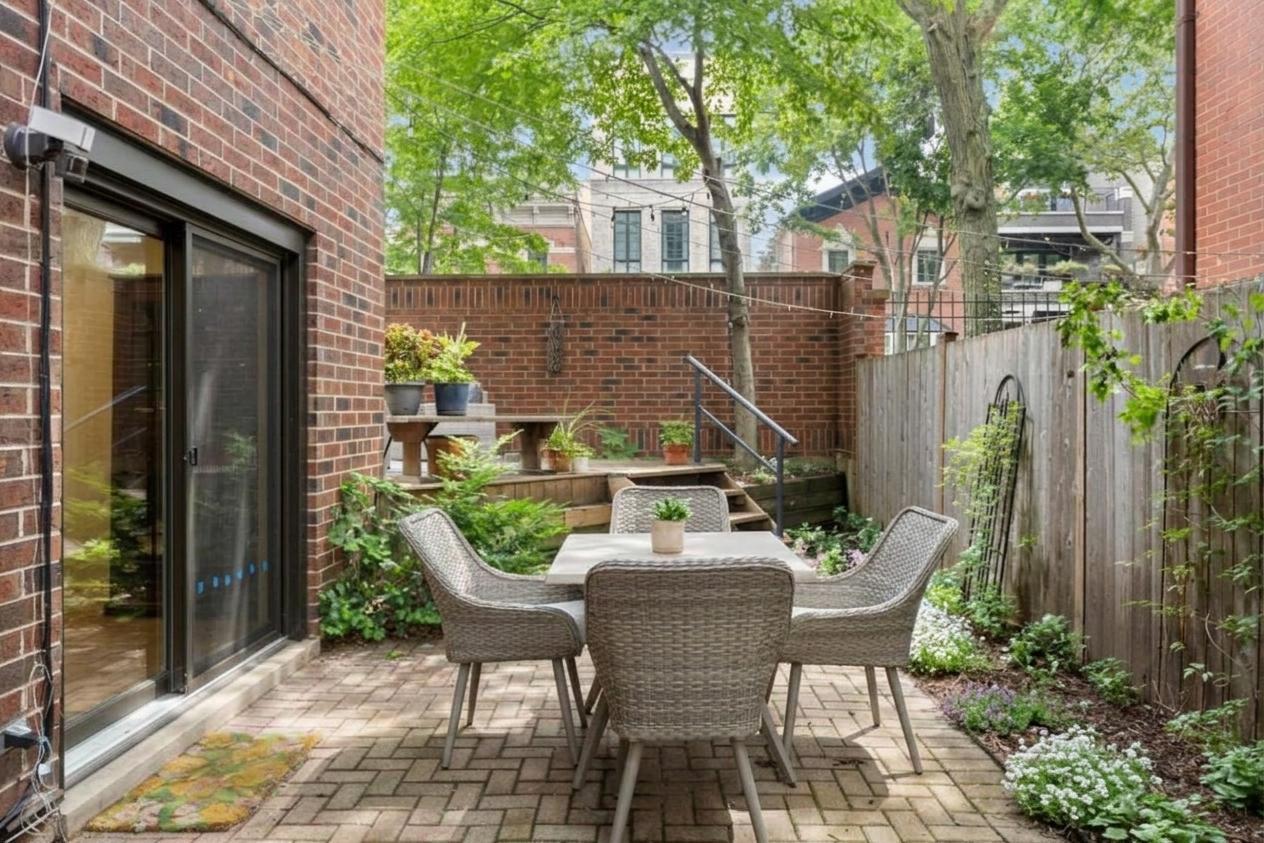 Brick patio with a wicker dining set, surrounded by leafy plants and a wooden fence.