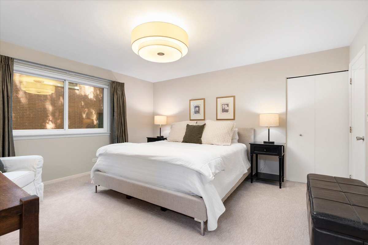 Staged bedroom with a white bed, bedside tables, wide curtained window, and bench.