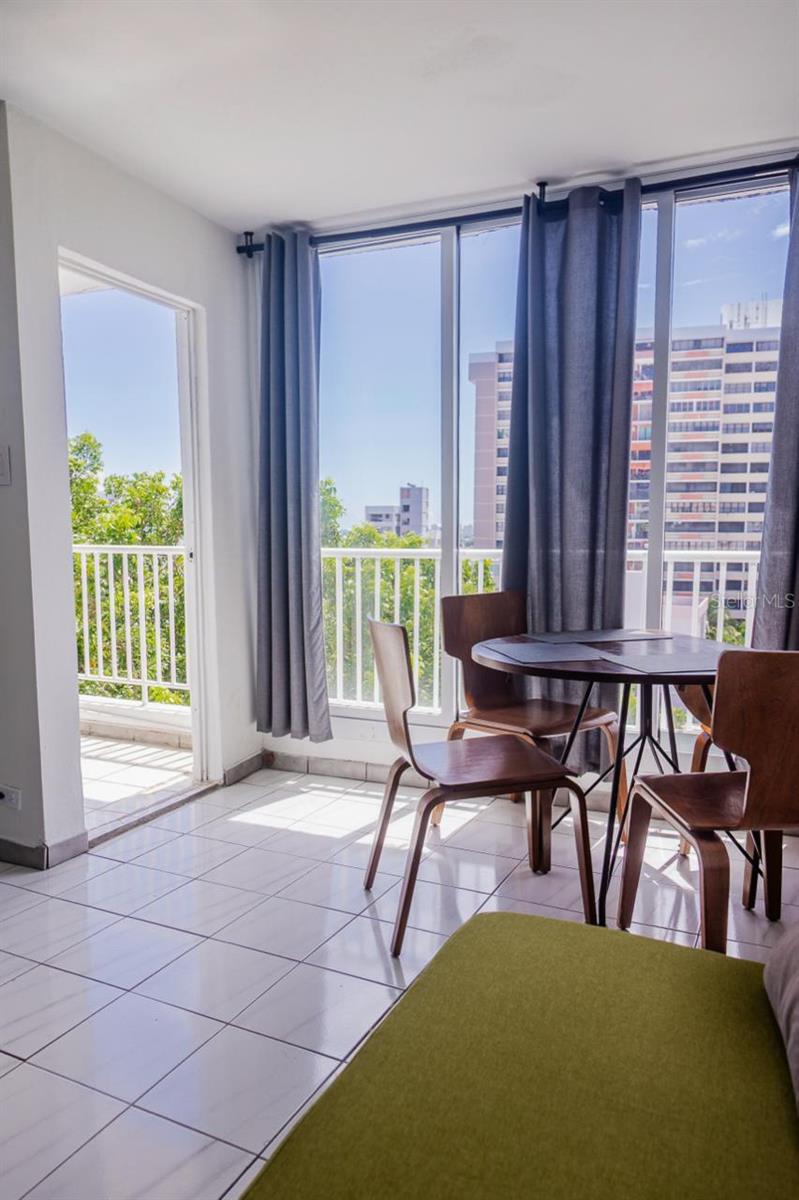 Dining area with a round wooden table beside floor-to-ceiling windows overlooking neighboring high-rises.