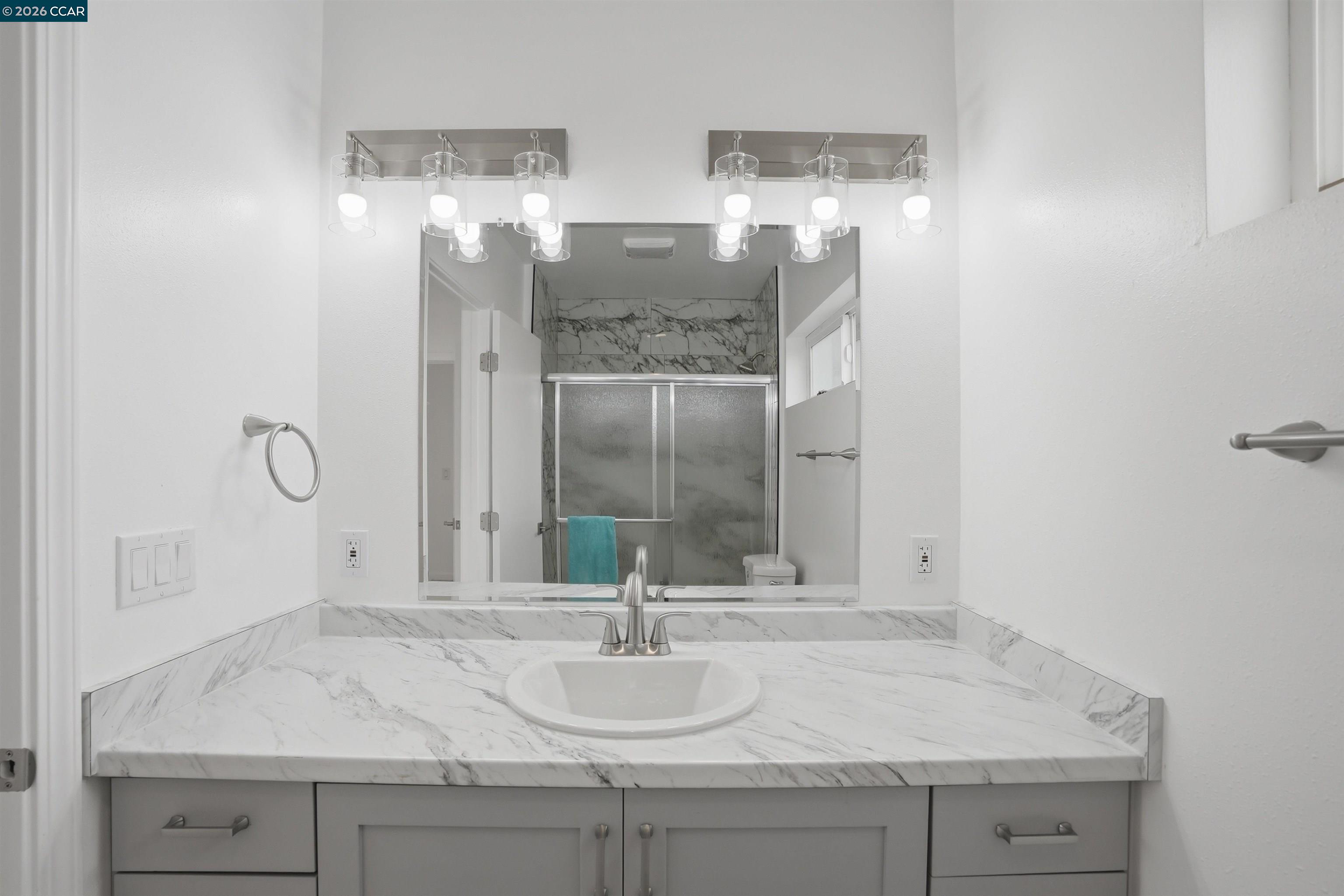 Bathroom vanity with a marble countertop, large mirror, and glass shower reflected behind it.