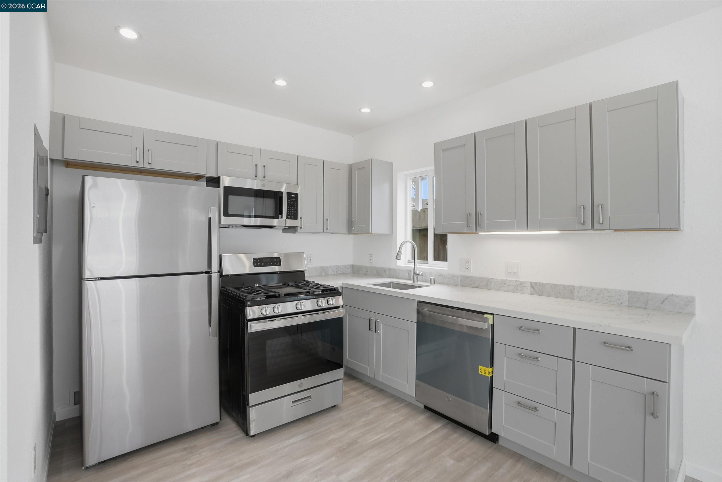 Kitchen with gray cabinets, stainless steel appliances, white counters, and a sink beneath the window.