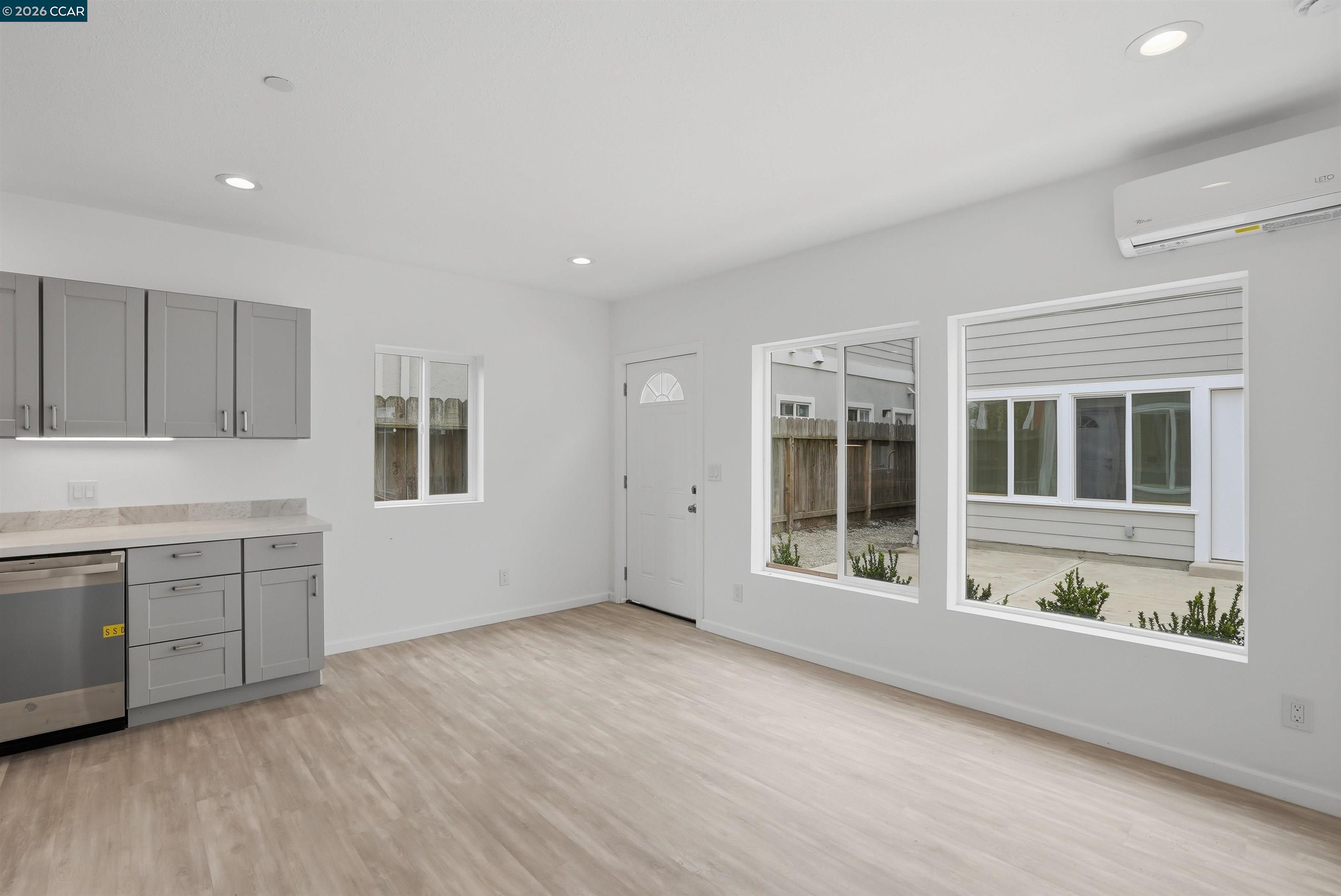 Vacant kitchen and living area with gray cabinets, light wood-look floors, large windows, and a wall-mounted mini-split.