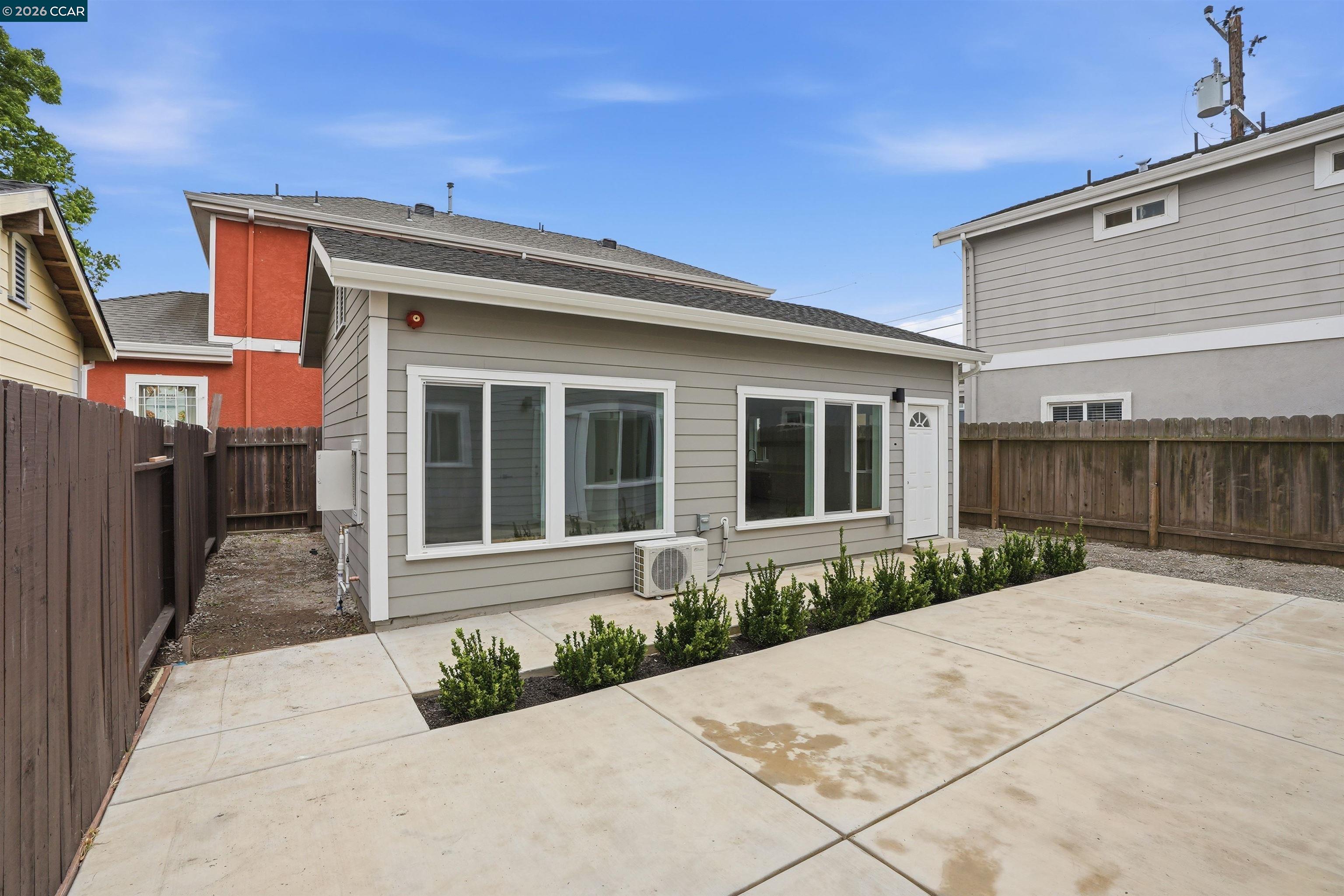 Rear of a gray-sided home with a broad concrete patio, fenced yard, and foundation shrubs.