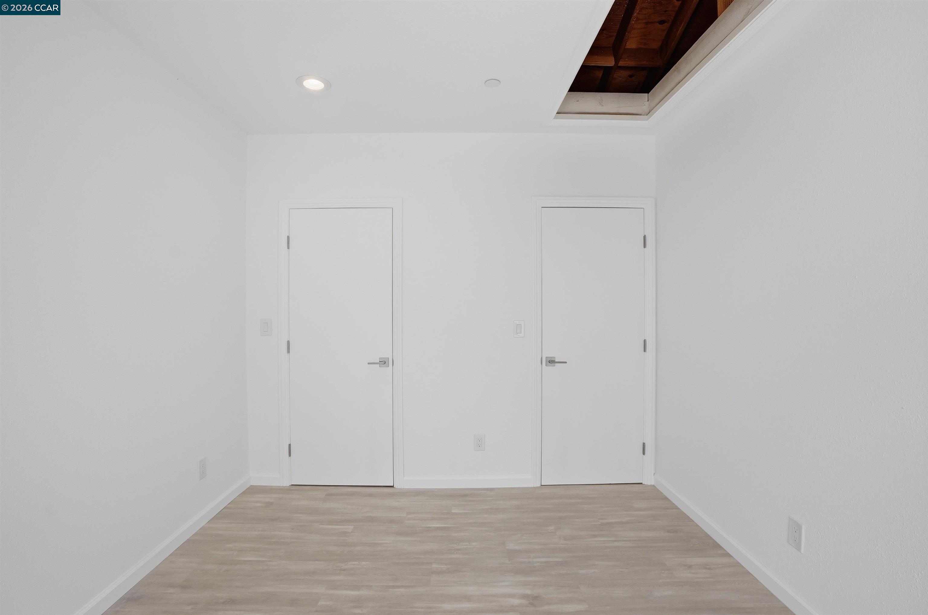 Empty white room with pale wood-look flooring, two doors, and an exposed wood ceiling inset.