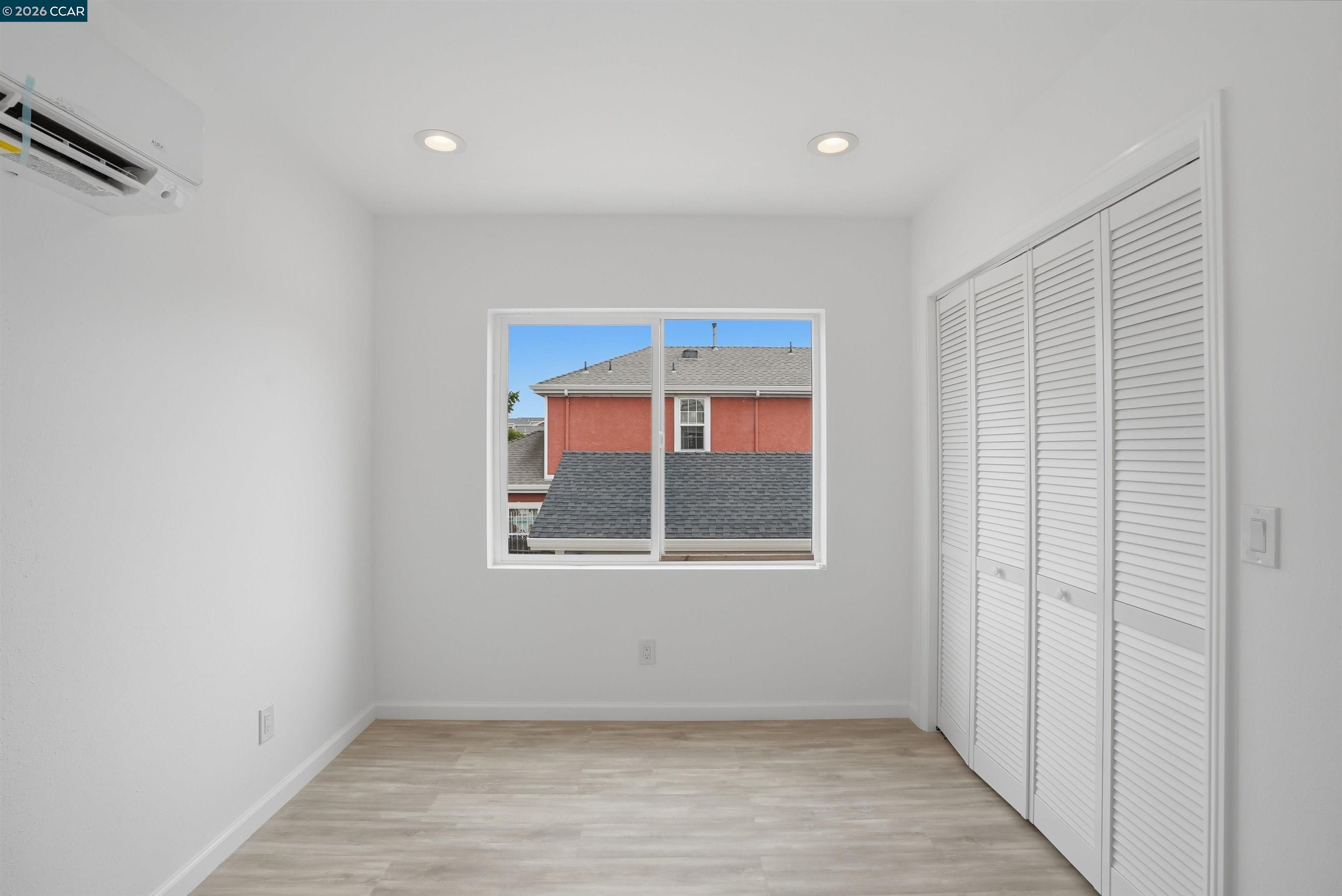 Empty room with louvered closets, a mini-split air conditioner, and a window overlooking a neighboring building.