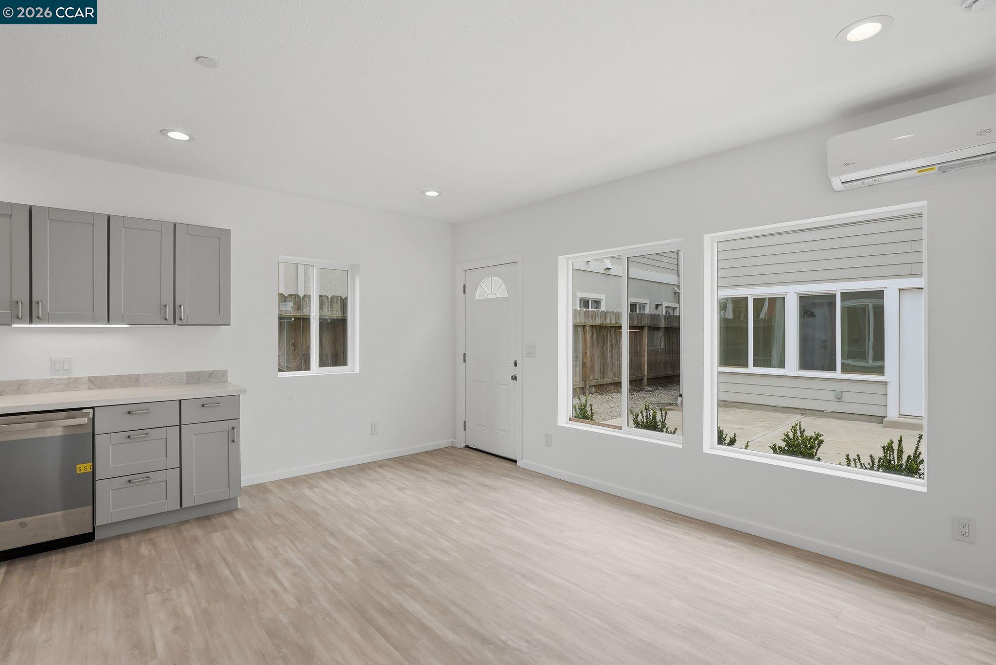Vacant kitchen and living area with gray cabinets, light flooring, and large windows overlooking a fenced patio.