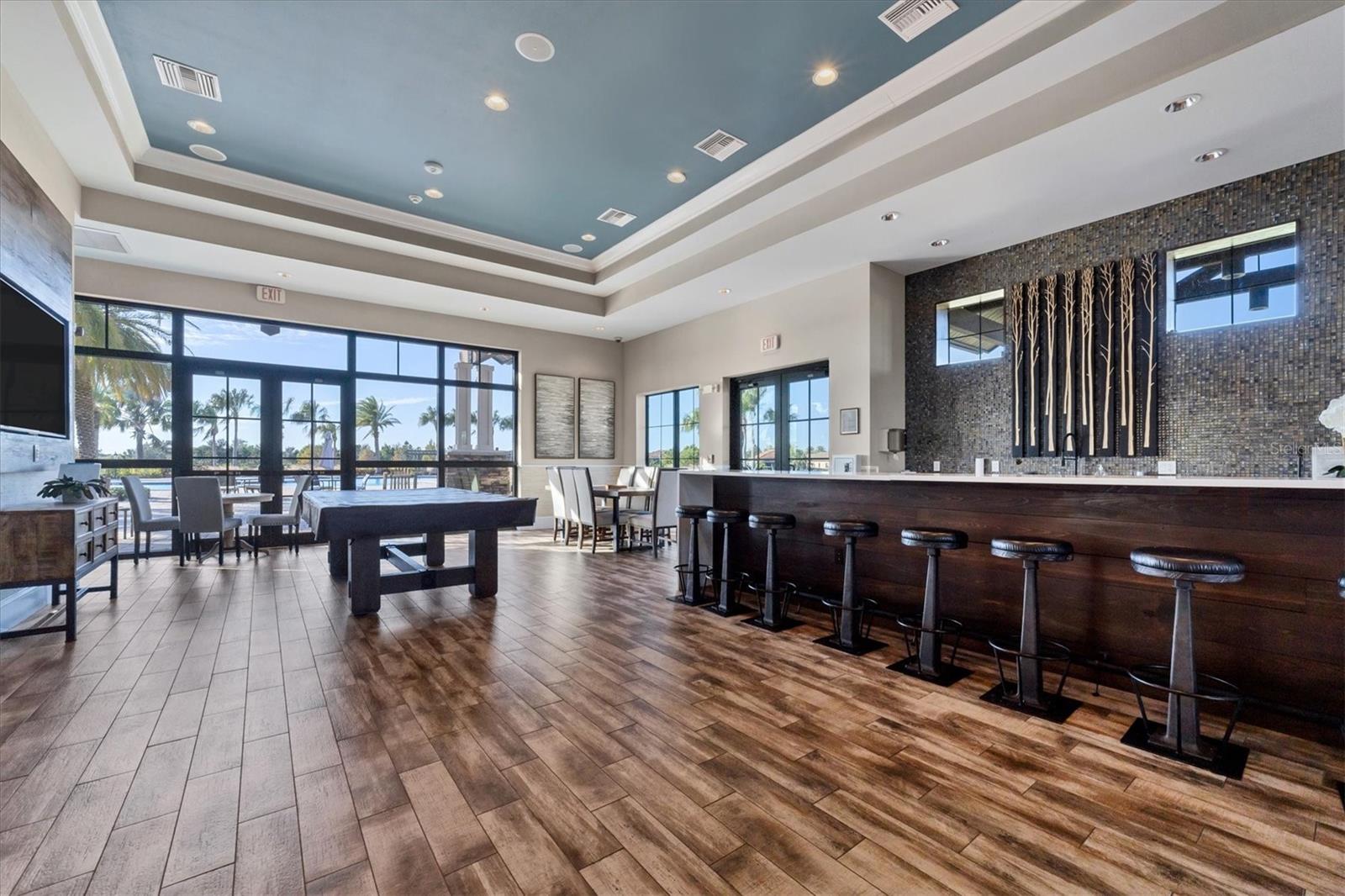 Furnished lounge with a long bar, game table, and windows overlooking a pool.