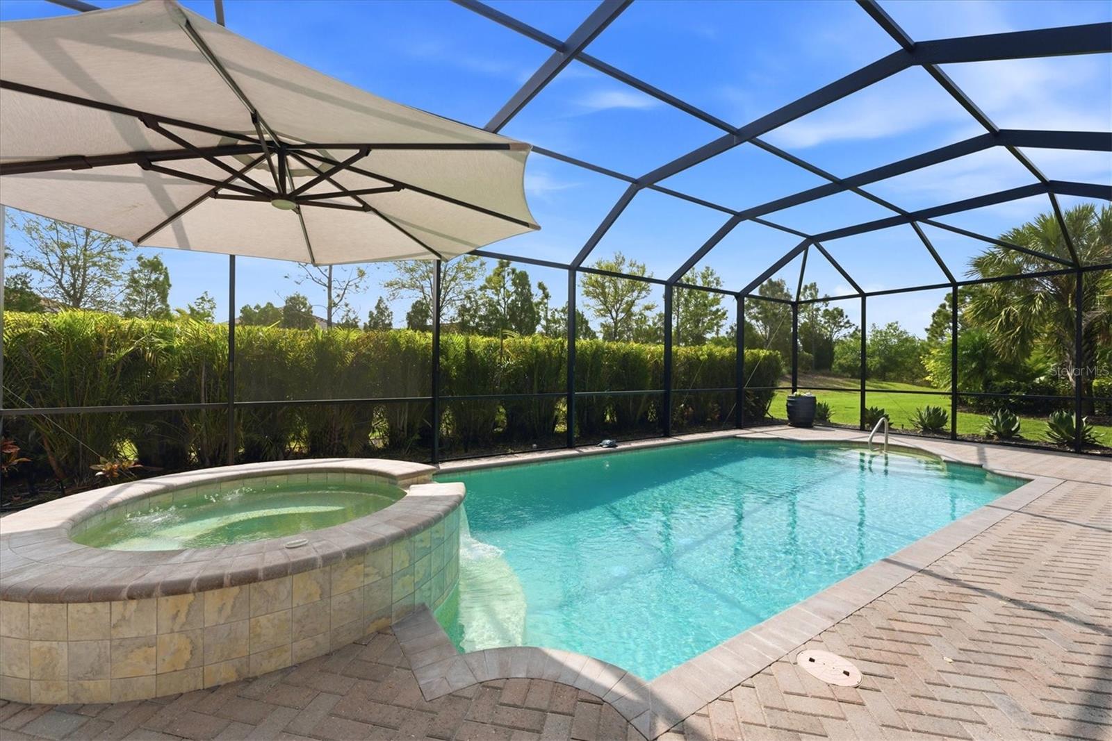 Screened backyard pool and raised spa beside a paved patio, shaded by a large umbrella.