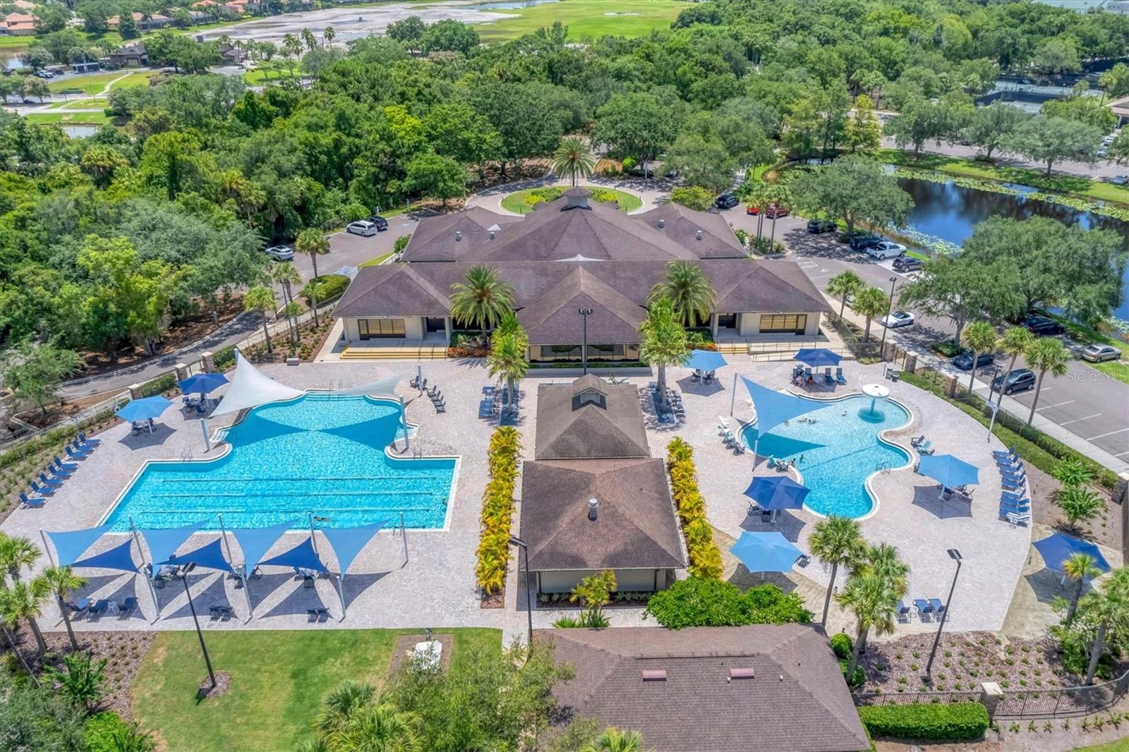 Aerial view of a clubhouse between two pools, with blue shade umbrellas and a pond nearby.