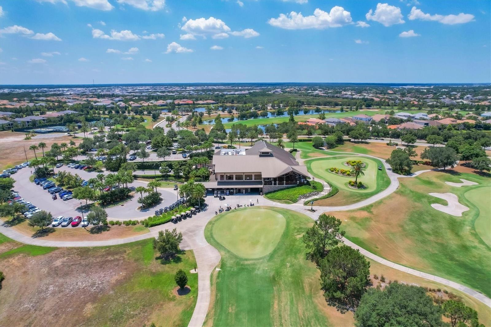 Aerial view of a golf course clubhouse and putting greens, with a lake and homes in the distance.