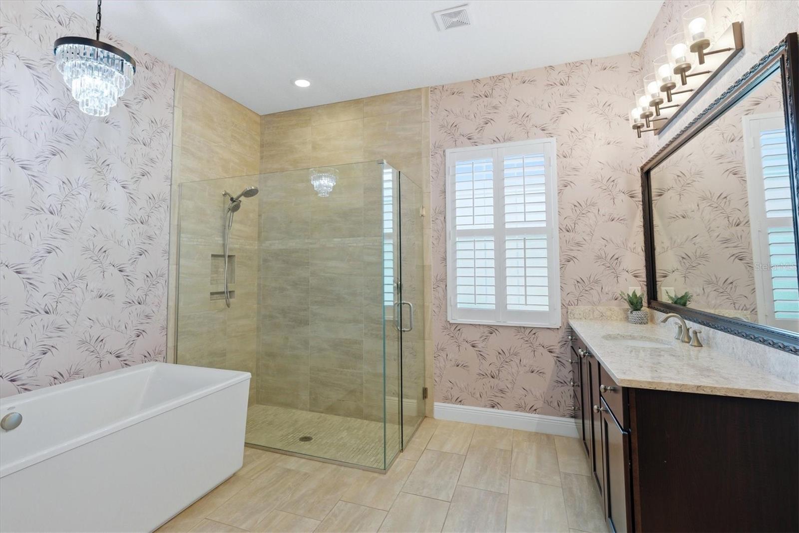 Bathroom with a freestanding tub, tiled glass shower, patterned wallpaper, and dark double vanity.
