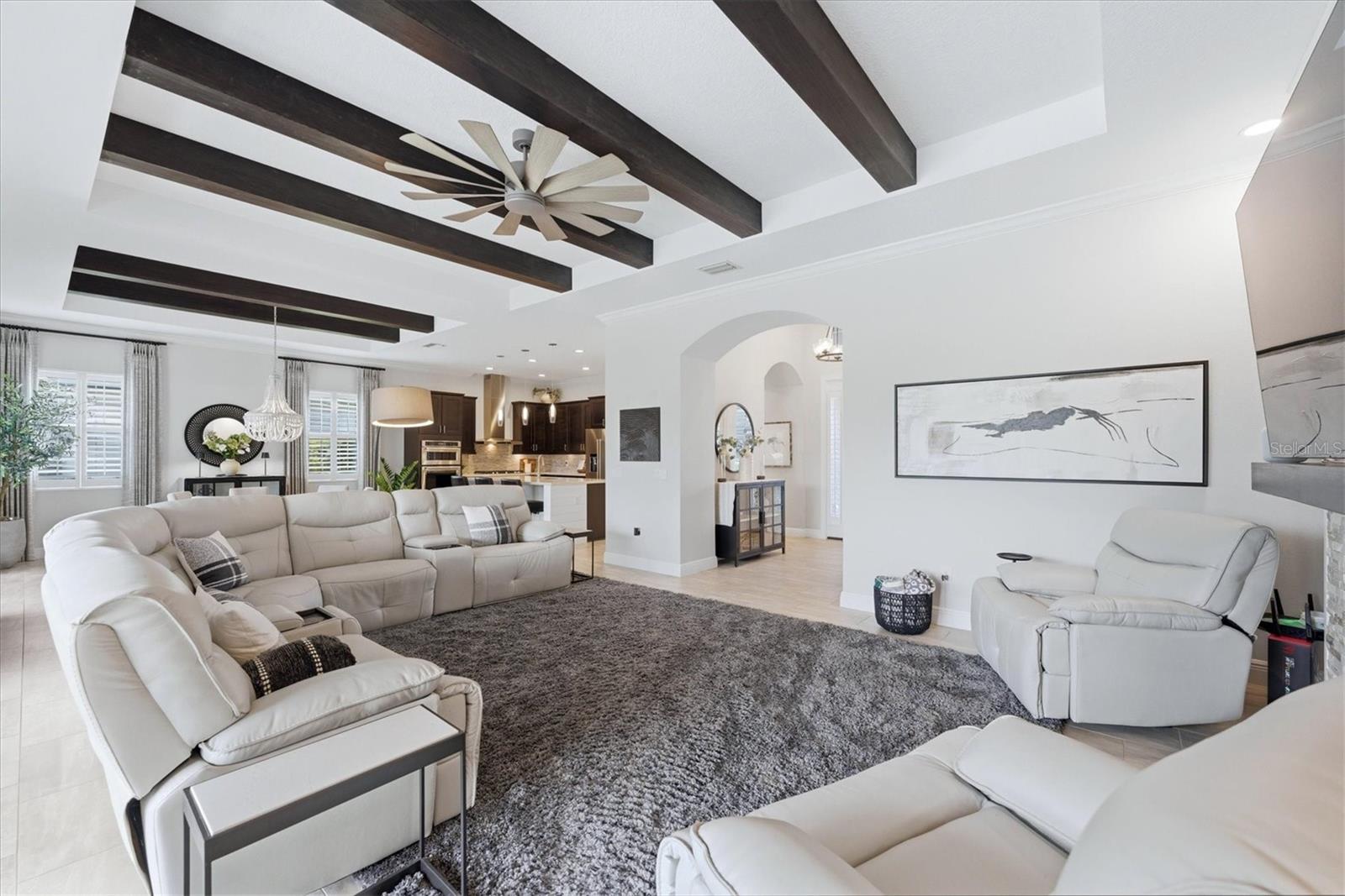 Living room with white sectional seating, dark ceiling beams, and an open kitchen
