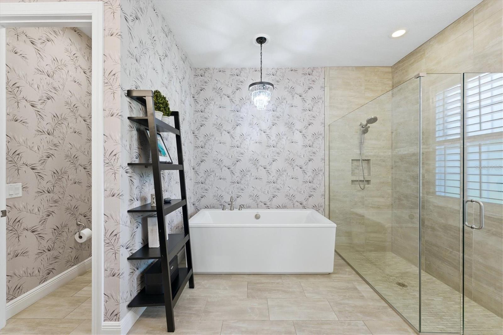 Bathroom with a freestanding soaking tub, glass shower, botanical wallpaper, and crystal chandelier.