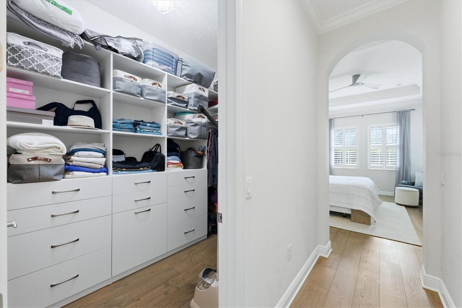 Walk-in closet with built-in white drawers and shelves opens toward a bedroom.