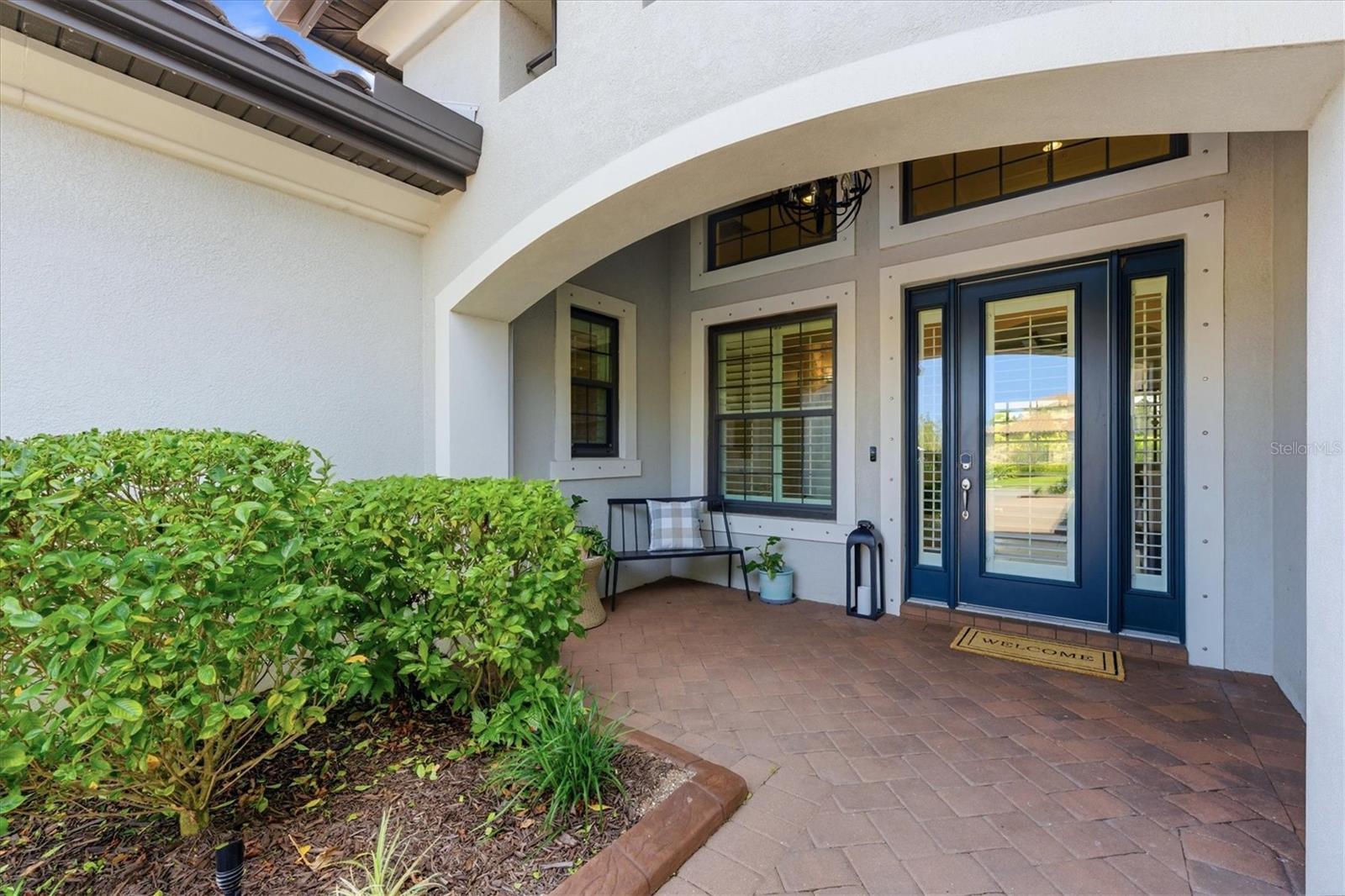 Home entrance with an arched stucco porch, blue double doors, brick pavers, and trimmed shrubs.