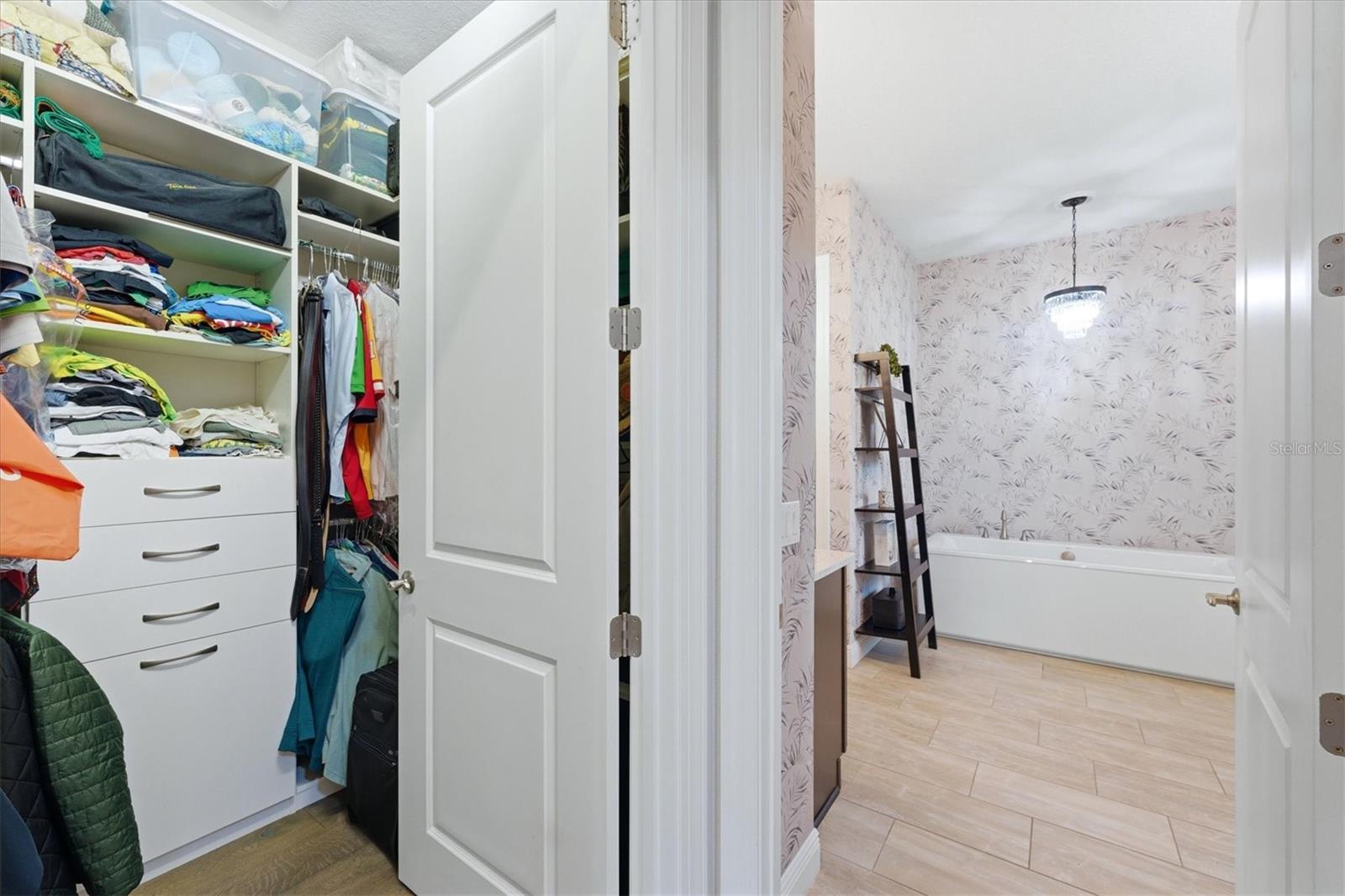 Walk-in closet opening into a bathroom with a bathtub and patterned wallpaper.