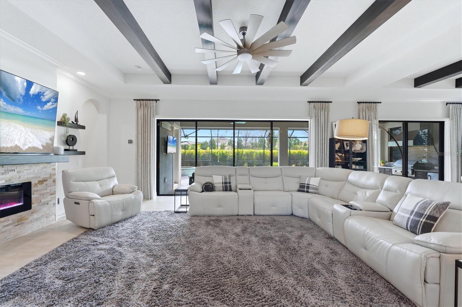 Living room with a white sectional, gray rug, stone fireplace, and dark ceiling beams.