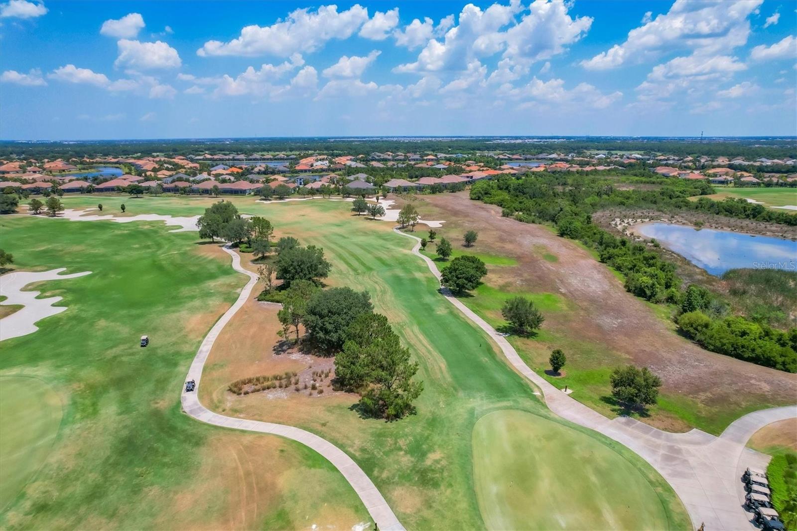 Aerial golf course with winding cart paths, bunkers, a pond, and homes in the distance.