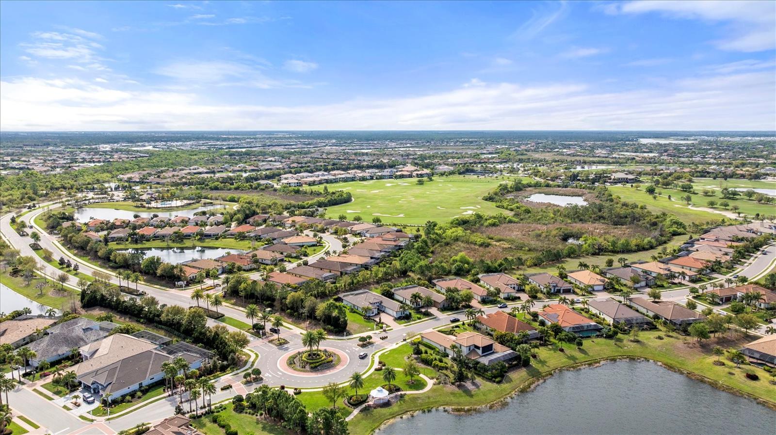 Aerial view of homes around a golf course and lake, with a landscaped roundabout in the foreground.